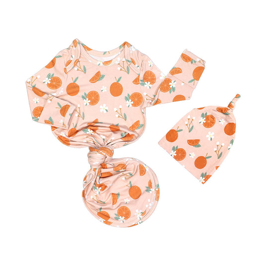the "freshly squeezed" bamboo gown and hat newborn set. the bottom half of the gown is tied in a knot. the "freshly squeezed" print has an assortment of full and half cut oranges scattered around. there is also flower heads and flower stems that intermingle within the print. this is all space out around a pink background space. 