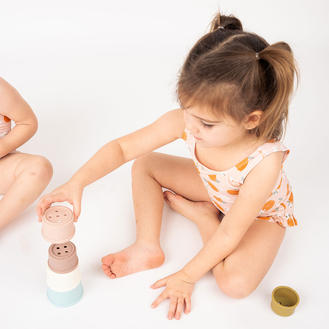 little girl playing with the silicone cups. she is wearing her "freshly squeezed" one piece bathing suit. the "freshly squeezed" print has an assortment of full and half cut oranges scattered around. there is also flower heads and flower stems that intermingle within the print. this is all space out around a pink background space. 