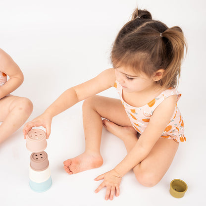 little girl playing with the silicone cups. she is wearing her "freshly squeezed" one piece bathing suit. the "freshly squeezed" print has an assortment of full and half cut oranges scattered around. there is also flower heads and flower stems that intermingle within the print. this is all space out around a pink background space. 