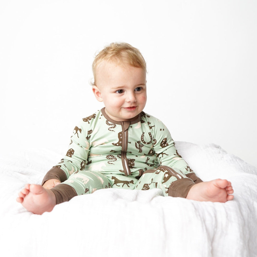 a small baby sits and smiles on a white blanket. the baby wears the "giddy up" convertible. the "giddy up" print is a western look at horses, cowboy hats, boots, horse rings, cacti, moons and stars. colors on this print include green, brown, and white. 