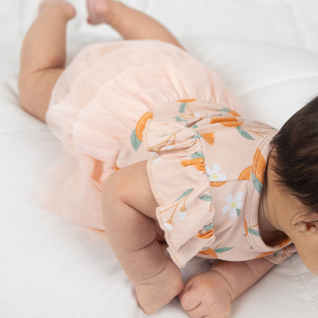 a zoomed in photo of a baby play on its tummy, wearing the "freshly squeezed" skirted onesie. this picture highlights the fact that the skirt wraps all the way around the onesie. the "freshly squeezed" print has an assortment of full and half cut oranges scattered around. there is also flower heads and flower stems that intermingle within the print. this is all space out around a pink background space. 