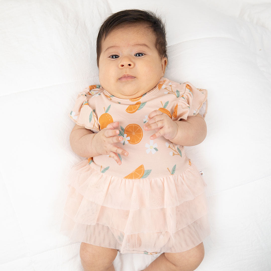 a baby is laying on its back in the "freshly squeezed" skirted onesie. the "freshly squeezed" print has an assortment of full and half cut oranges scattered around. there is also flower heads and flower stems that intermingle within the print. this is all space out around a pink background space. 