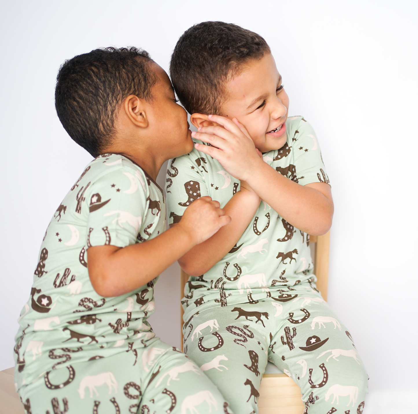 2 boys sit next to one another. one boy whispers something into the others ear. they are both wearing the "giddy up" matching pajama set. the "giddy up" print is a western look at horses, cowboy hats, boots, horse rings, cacti, moons and stars. colors on this print include green, brown, and white. 