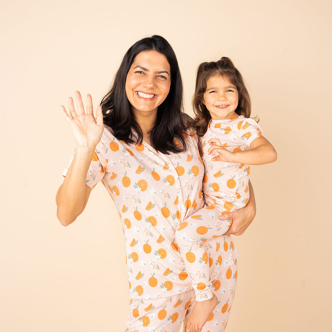 mother and her toddler waving, they are both wearing "freshly squeezed" prints. the mom is in the women's top and the joggers. the toddler is in the 2-piece matching pajama set. the "freshly squeezed" print has an assortment of full and half cut oranges scattered around. there is also flower heads and flower stems that intermingle within the print. this is all space out around a pink background space. 