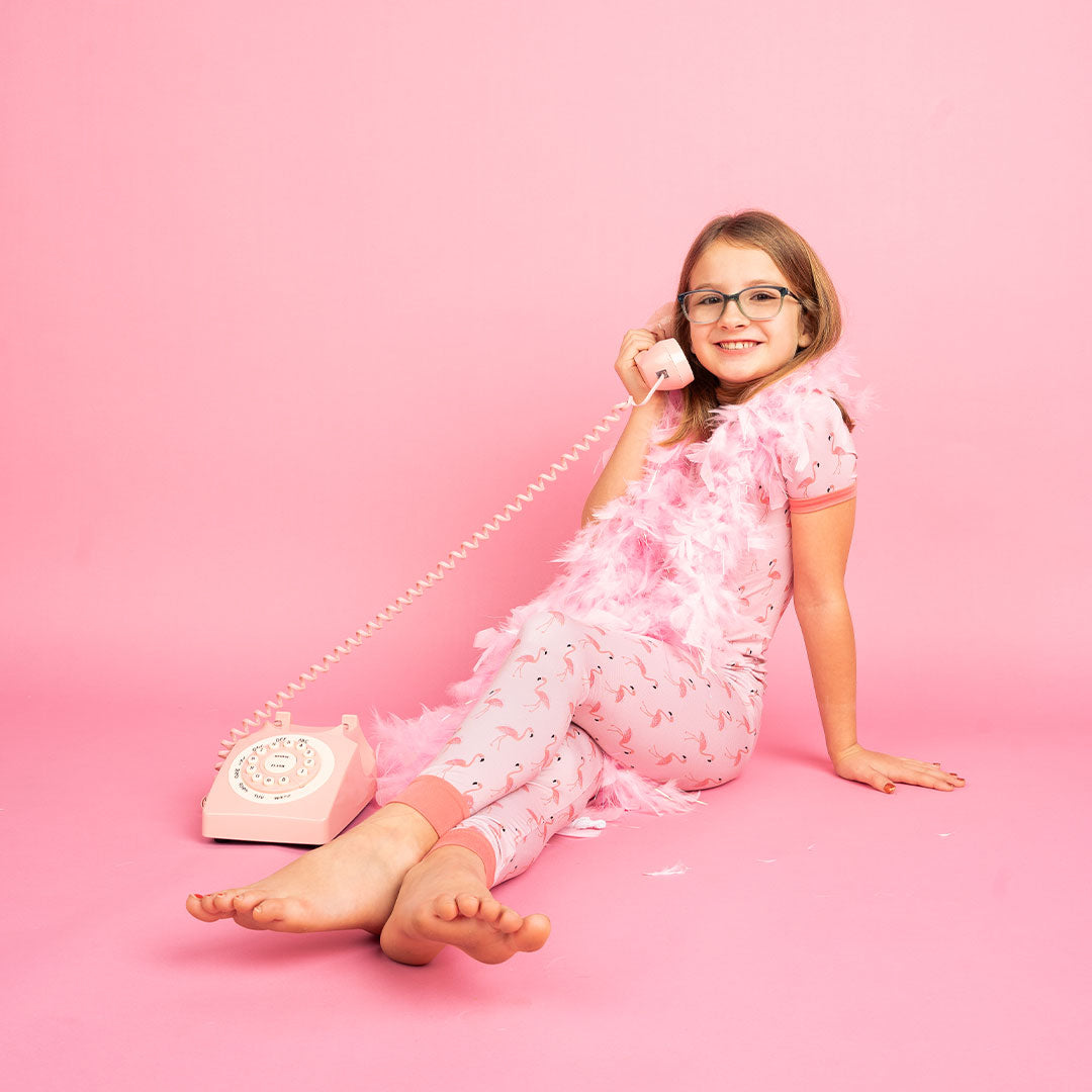 young girl in her matching 2-piece "fancy flamingos" pajama set. she is wearing a pink feathered boa scarf and playing with an old-school telephone. the "fancy flamingos" print is a pattern of multiple pink flamingoes scattered around the print. 