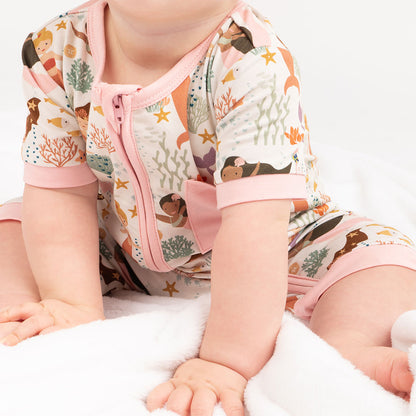 an up close, focused picture of a baby sitting on the floor. she is wearing the "making waves" shortie. the "making waves" print has a diverse spread of mermaids, sea coral, starfish, fish, and bubbles all spread out in different colors. this is all put on a beige background.