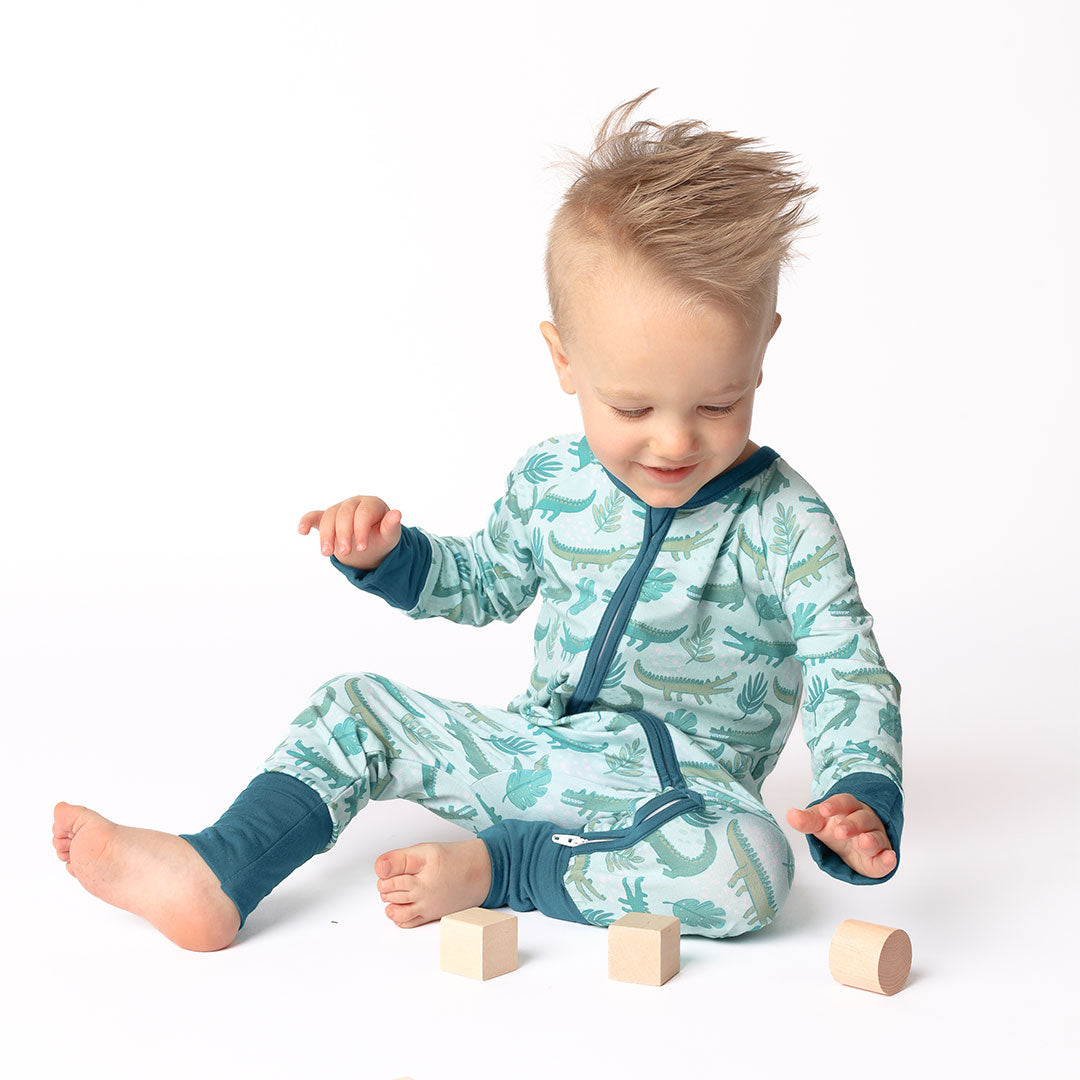 Little toddler boy playing with blocks. He is wearing his "later gator" convertible. the "later gator" print has a mix of light and dark green alligators, leaves, and white dots scattered on a teal colored background. 