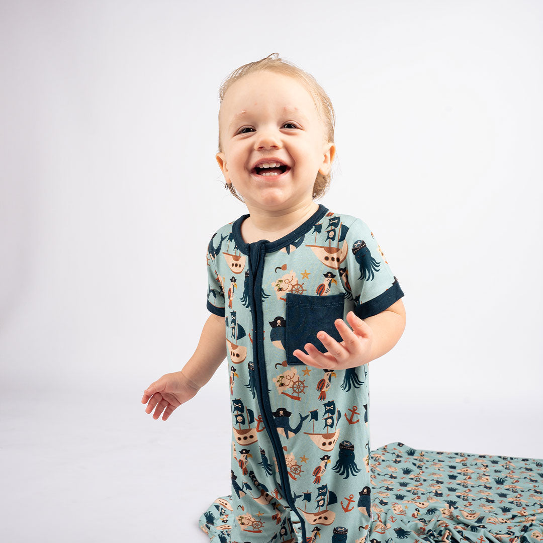 baby laughing at the camera in his "pirate's life" shortie. you can see the "pirate's life" bamboo blanket behind him. the "pirate's life" print is a mix of octopus, whales, and seagulls all wearing pirate hats. there are also anchors, pirate ships, treasure maps, and starfish mixed into the pattern. this is all displayed on a teal background. 