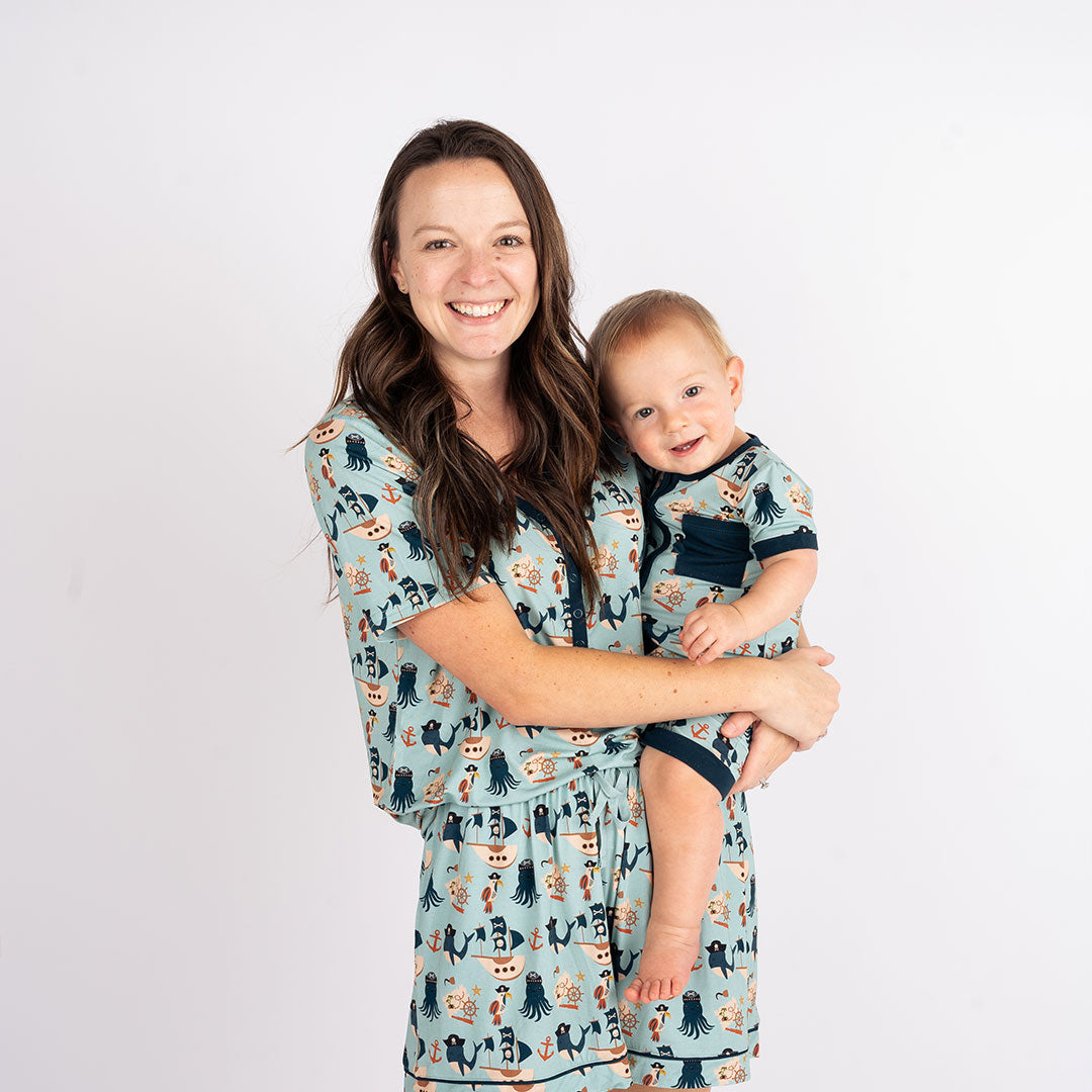 a mom smiles at the camera with her baby smiling on her hip. they are both wearing matching "pirate's life" prints. mom is wearing the women's top and women's shorts. the baby is in the shhortie. the "pirate's life" print is a mix of octopus, whales, and seagulls all wearing pirate hats. there are also anchors, pirate ships, treasure maps, and starfish mixed into the pattern. this is all displayed on a teal background. 