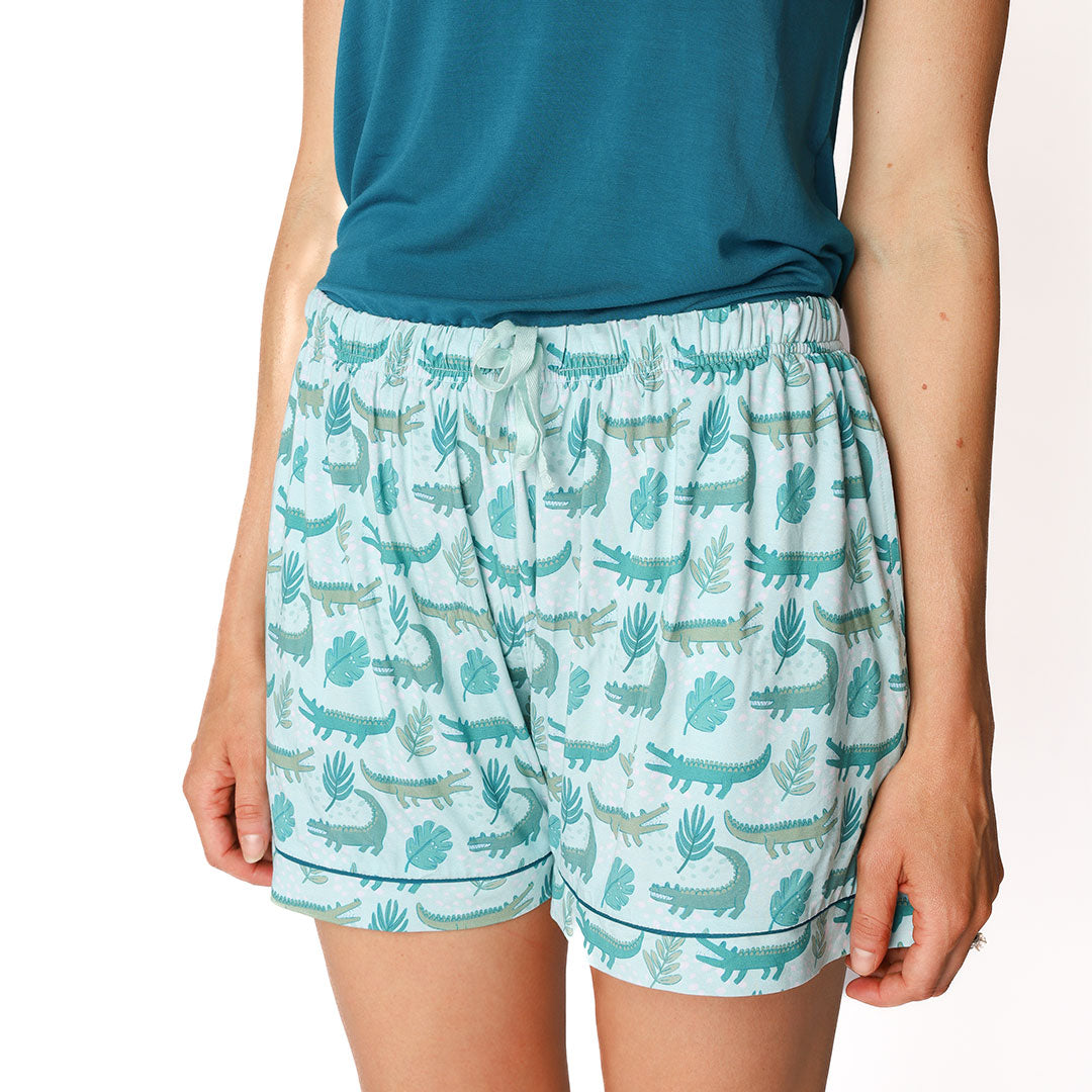 woman posing in her "later gator" women's bamboo pajama shorts. the "later gator" print has a mix of light and dark green alligators, leaves, and white dots scattered on a teal colored background. 