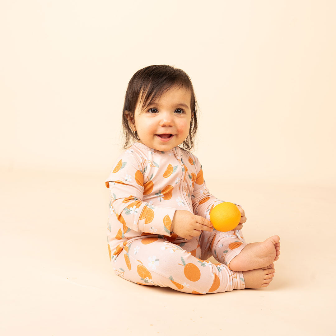 a happy baby sitting on the floor with a orange in her hand. she is wearing the "freshly squeezed" convertible. the "freshly squeezed" print has an assortment of full and half cut oranges scattered around. there is also flower heads and flower stems that intermingle within the print. this is all space out around a pink background space. 