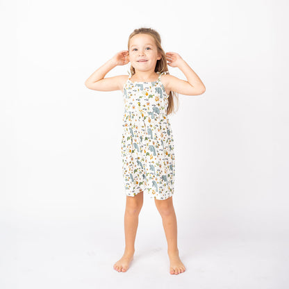 a girl stands with her hands behind her head. she smiles wide while wearing her "manatee" sundress. the print "manatee" unites the florida sea life in a majestic print. there is a variation of florida manatees, yellow and pink sea horses, yellow and pink starfish, bubbles, turtles, and coral patterned across a white background