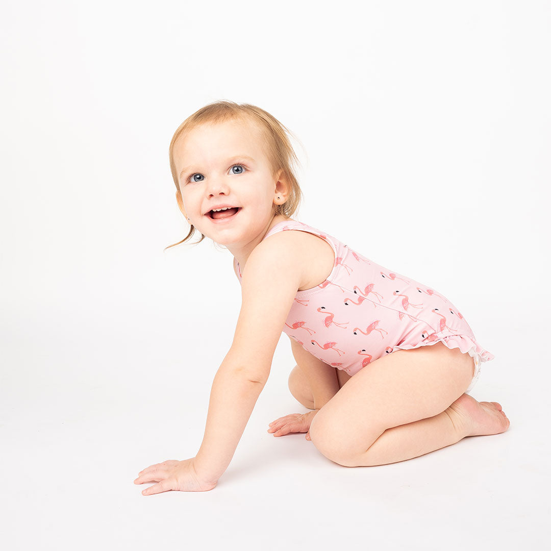 a baby girl crawling on the floor in her "fancy flamingos" one piece bathing suit. the "fancy flamingos" print is a pattern of multiple pink flamingoes scattered around the print. 