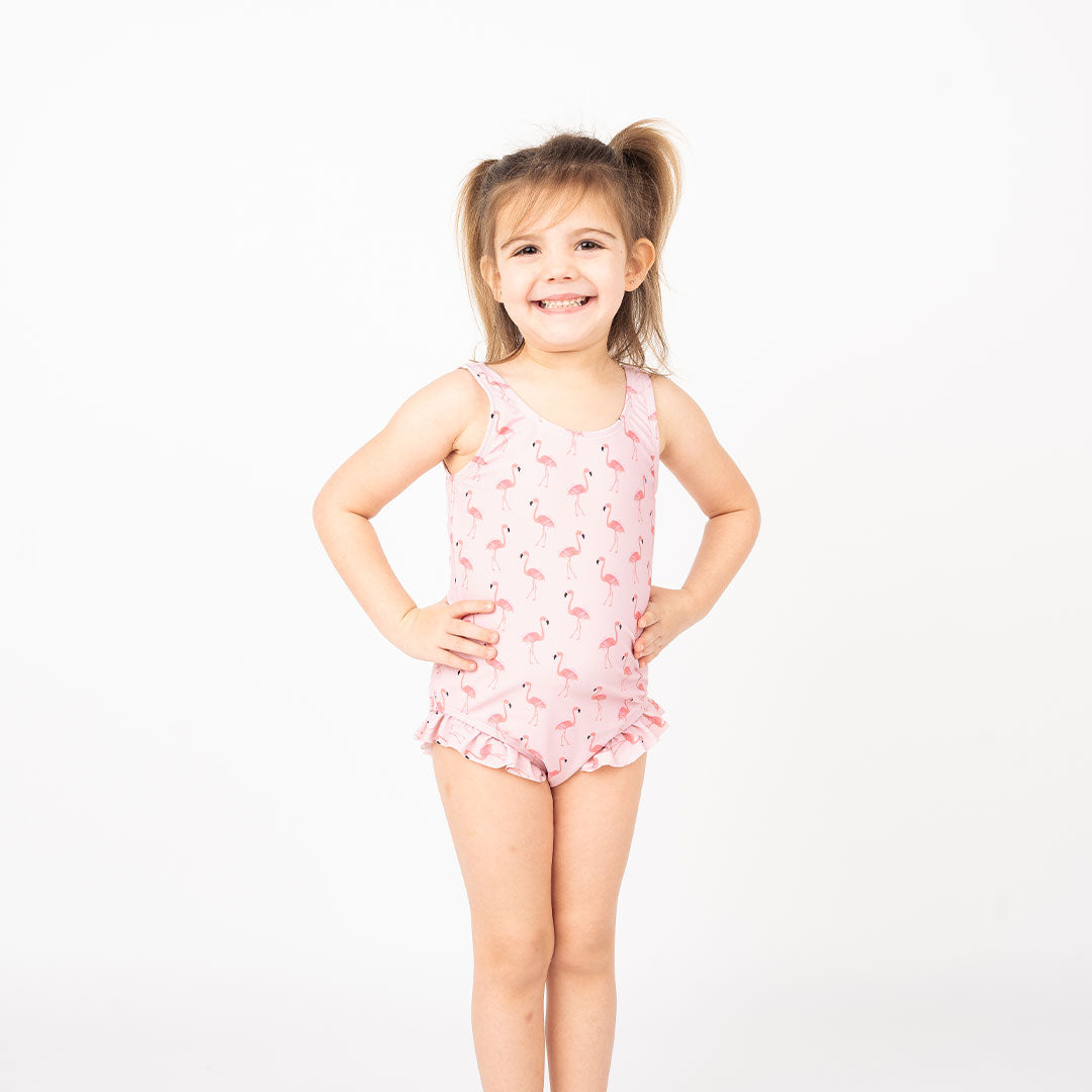 cute toddler girl smiling in her "fancy flamingos" one piece bathing suit. the "fancy flamingos" print is a pattern of multiple pink flamingoes scattered around the print. 