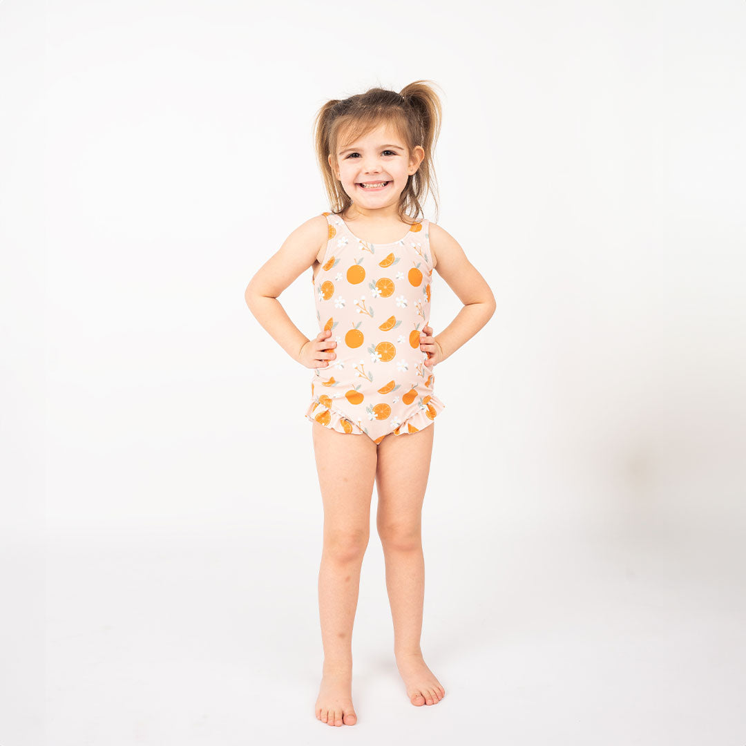 girl posing with her hands on her hips, smiling big, wearing her "freshly squeezed" one piece swim suit. the "freshly squeezed" print has an assortment of full and half cut oranges scattered around. there is also flower heads and flower stems that intermingle within the print. this is all space out around a pink background space. 