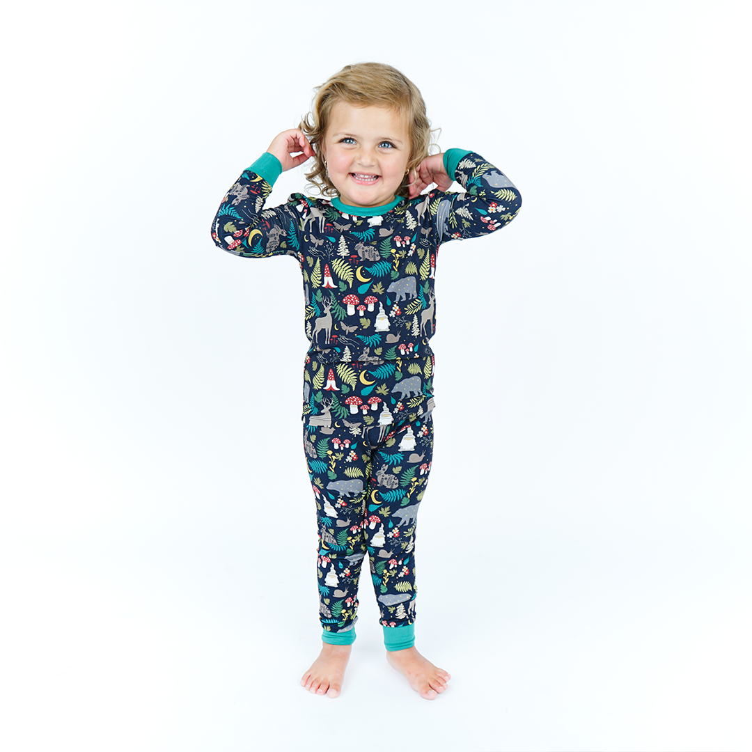 a young girl smiles with her hands up, she is wearing the "night forest" 2-piece matching pajama set. the "night forest" print is a night time themed design. you can see an array of forest animals ranging from, deer, bears, bunnies, birds, forest trees and leaves, flowers, and mushrooms. there are also starts and moons scattered around to enhance the night time atmosphere. 