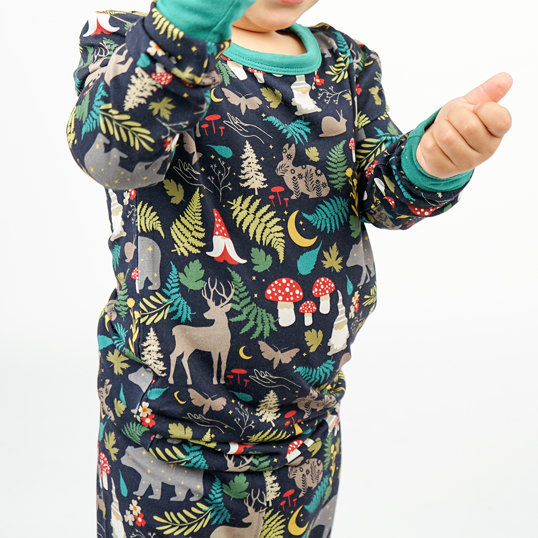 a close up picture of a child wearing the "night forest" 2-piece matching pajama set. the "night forest" print is a night time themed design. you can see an array of forest animals ranging from, deer, bears, bunnies, birds, forest trees and leaves, flowers, and mushrooms. there are also starts and moons scattered around to enhance the night time atmosphere. 