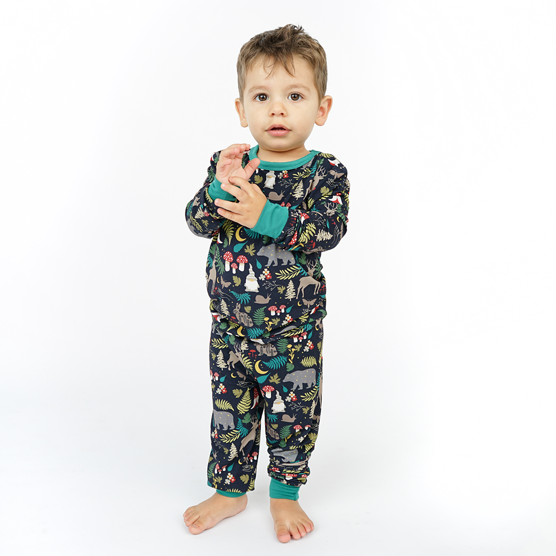 a young boy looks at the camera. he is standing in his "night forest" 2-piece matching pajama set. the "night forest" print is a night time themed design. you can see an array of forest animals ranging from, deer, bears, bunnies, birds, forest trees and leaves, flowers, and mushrooms. there are also starts and moons scattered around to enhance the night time atmosphere. 