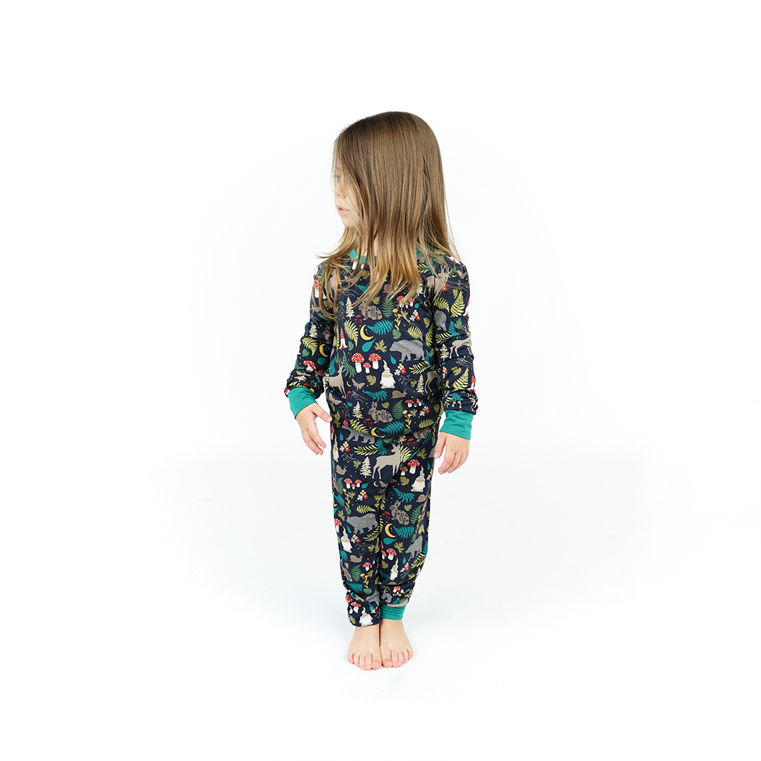 a young girl stands and looks to the left of her. she is wearing the "night forest" 2-piece pajama set. the "night forest" print is a night time themed design. you can see an array of forest animals ranging from, deer, bears, bunnies, birds, forest trees and leaves, flowers, and mushrooms. there are also starts and moons scattered around to enhance the night time atmosphere. 