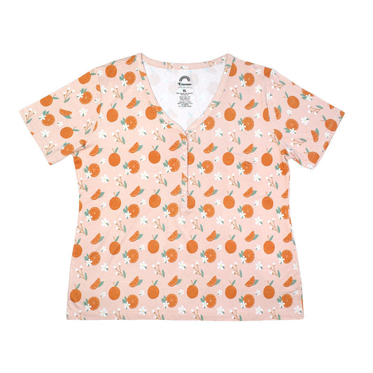 the "freshly squeezed" women's top. the "freshly squeezed" print has an assortment of full and half cut oranges scattered around. there is also flower heads and flower stems that intermingle within the print. this is all space out around a pink background space. 