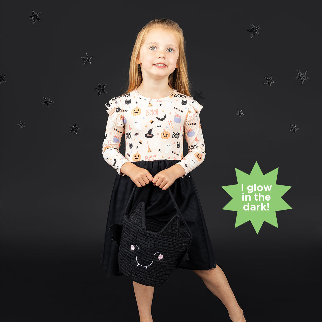 Little girl standing while smiling on black backdrop while holding black bat rope basket. She is modeling the tulle dress in  our Sweet and Spooky pink halloween Print featuring glow in the dark patterns, pumpkins, candy, bats and more!