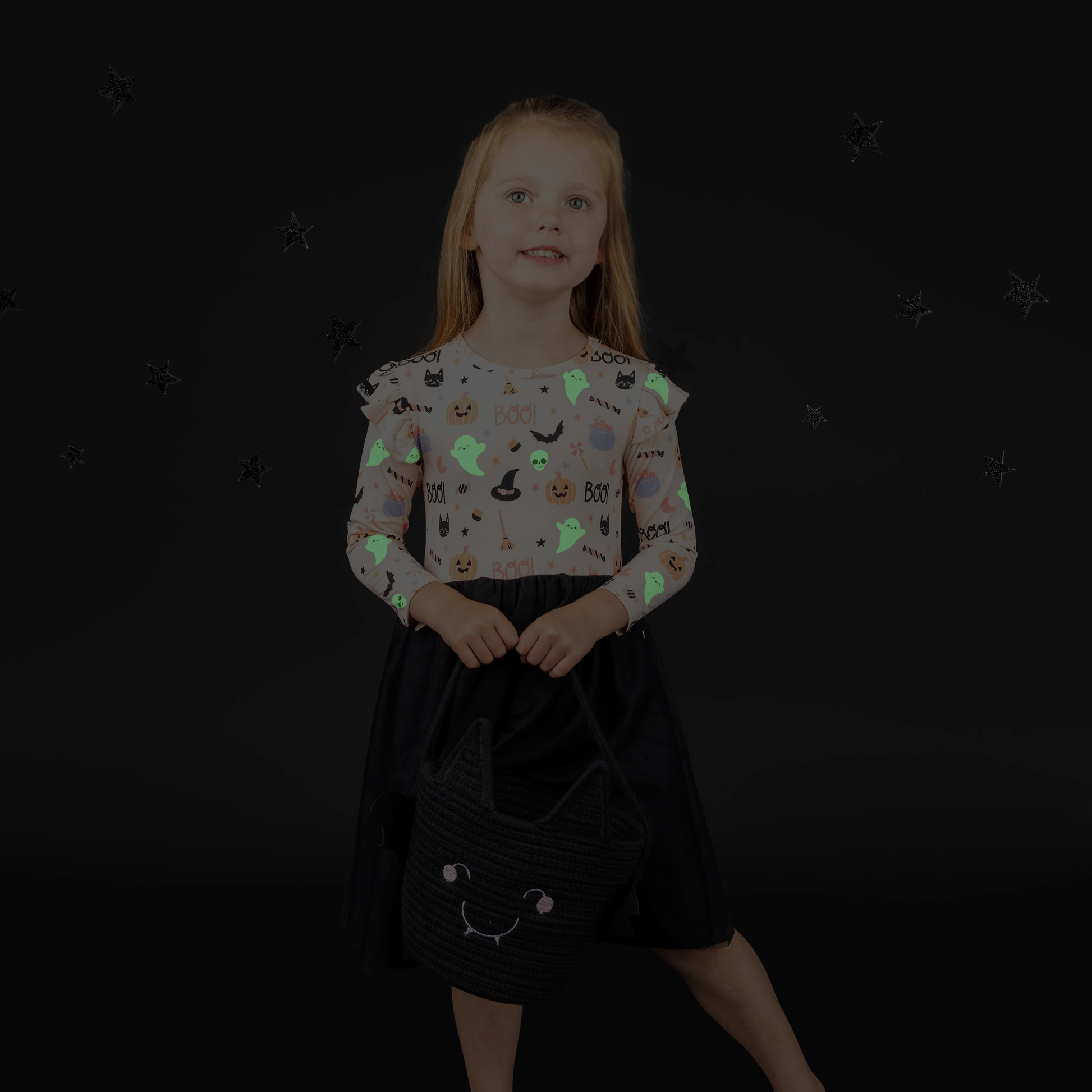 Little girl standing while smiling on black backdrop while holding black bat rope basket. She is modeling the tulle dress in  our Sweet and Spooky pink halloween Print featuring glow in the dark patterns, pumpkins, candy, bats and more!