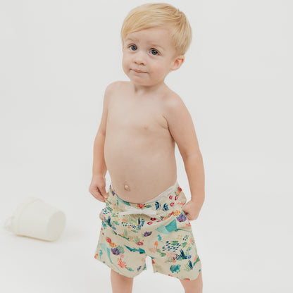 Toddler boy wearing our Emerson and Friends Coral Reef Boys Swim Trunks, featuring a vibrant ocean-themed design with sharks, sea turtles, and coral, made from quick-dry fabric with UPF sun protection for all-day beach and pool fun.