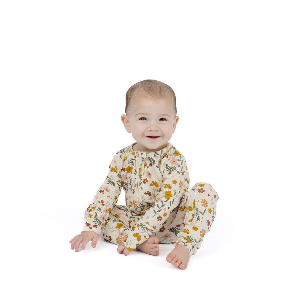 A baby sitting on a white studio background wearing a cream-colored long-sleeve footless onesie with a yellow and orange floral pattern.