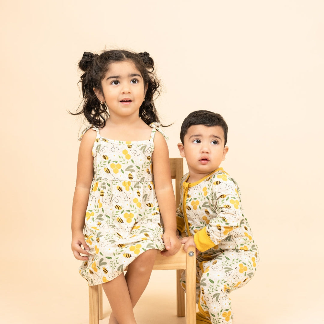 Toddler girl wearing a bamboo sundress with a honey bee print, sitting next to a baby boy in matching convertible bamboo pajamas. The soft, breathable fabric ensures comfort for both kids, perfect for spring and beyond.