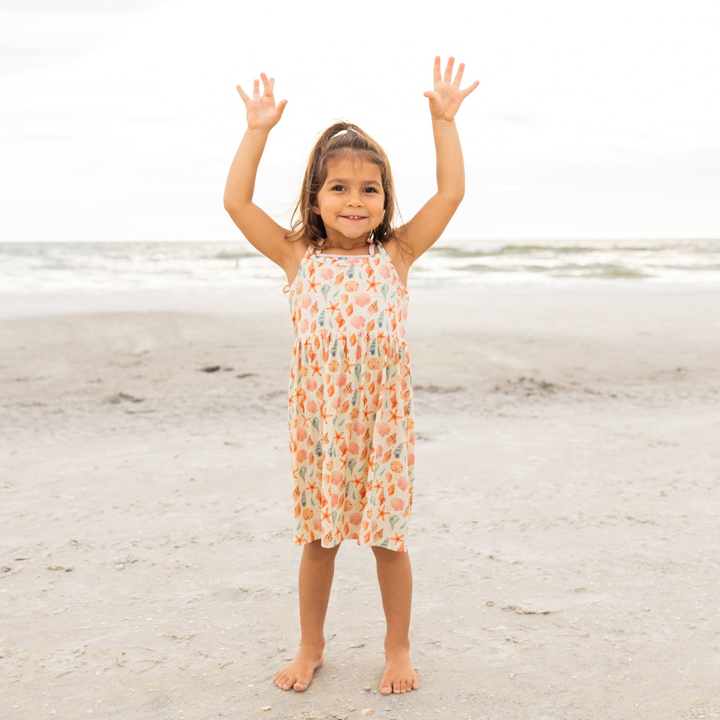 Little girl holding her hands up in the air on the beach wearing her Sandy Seashells Bamboo Sundress.