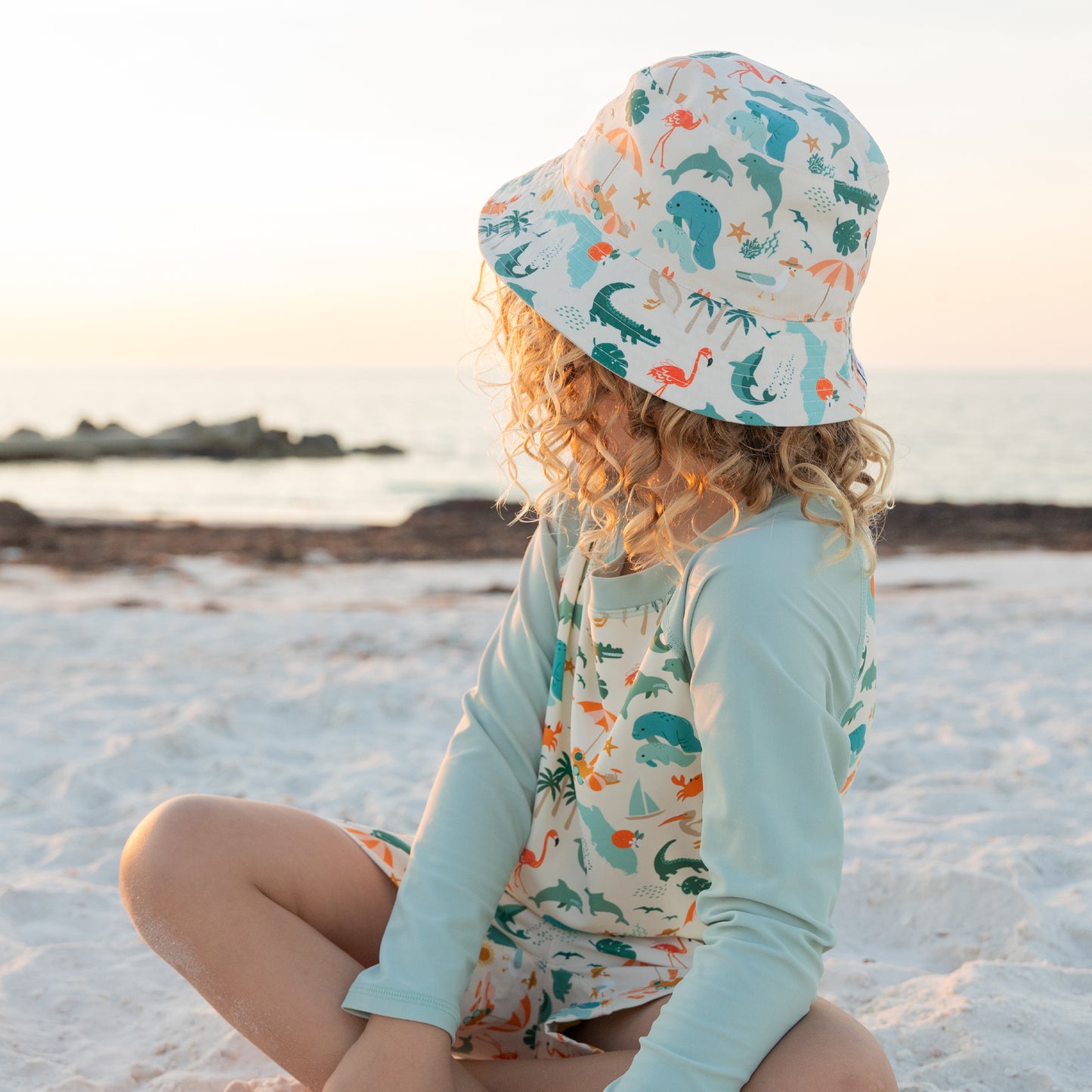 A young boy with curly hair sitting on the beach wearing the Explore Florida bucket hat and a matching long-sleeve rash guard.