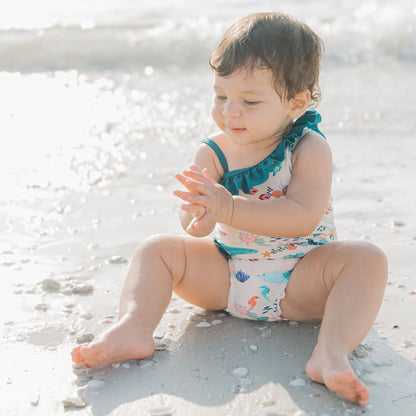 Toddler girl sitting at the beach wearing a Coral Reef Ruffle Leg One Piece Swimsuit with an ocean-themed print of sharks and sea turtles, designed with soft, stretchy fabric and UPF 50+ sun protection for a fun day at the beach.
