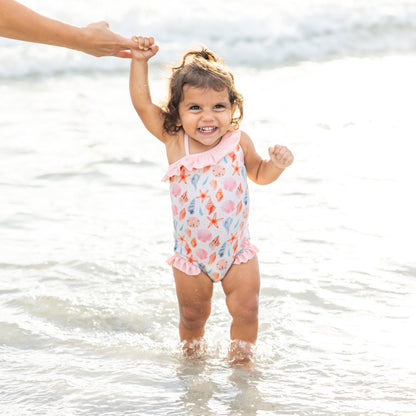 Little girl standing in the water wearing our  Sandy Seashells Ruffle Leg One Piece.