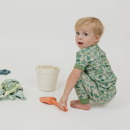 Toddler sitting in a white background playing with toys and wearing our Save the Sea Turtles bamboo short sleeve kids pajama pants set by Emerson and Friends. This ultra-soft, eco-friendly pajama set features an adorable sea turtle and ocean-inspired print, perfect for little nature lovers. Designed for all-night comfort with breathable bamboo fabric.
