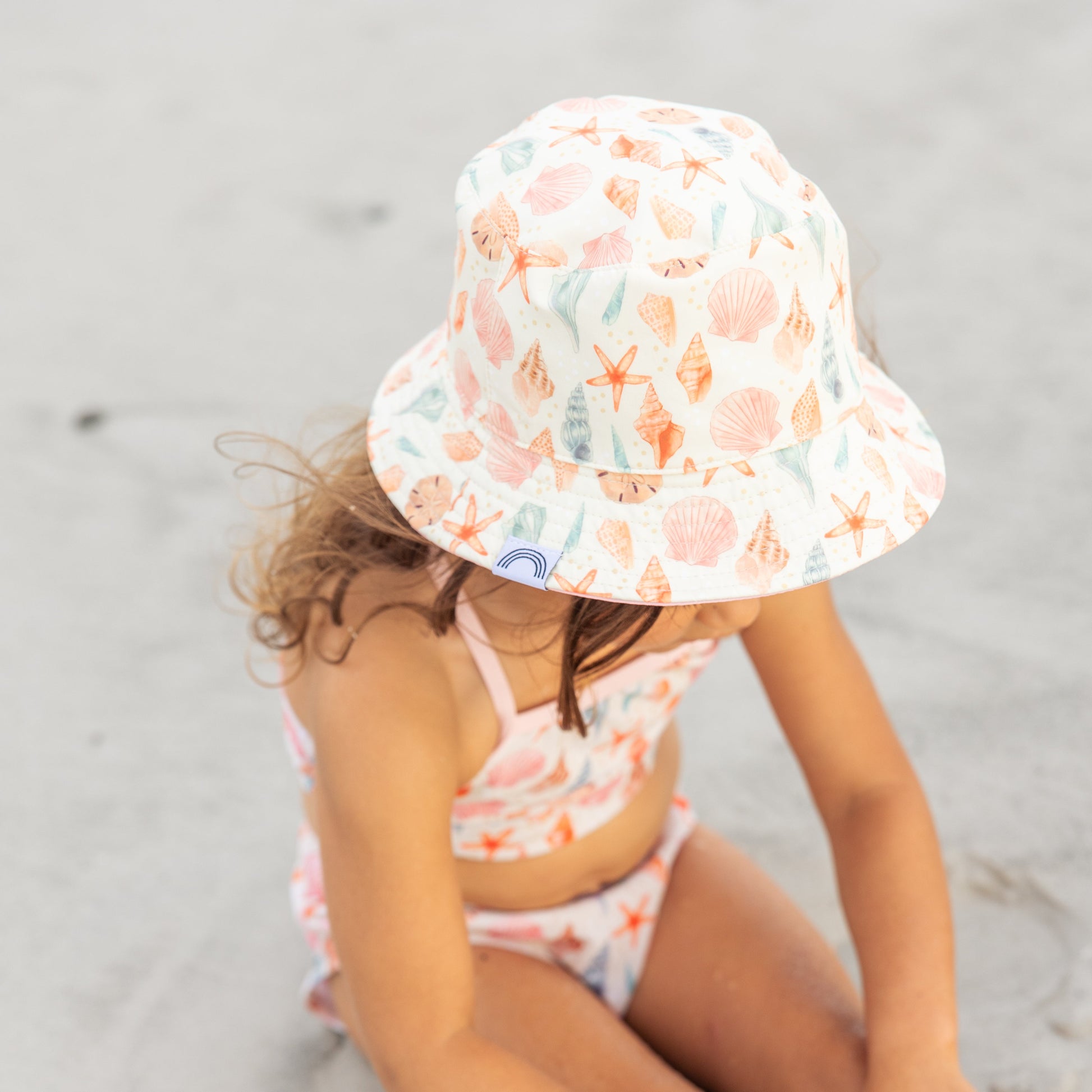 Little girl sitting in the sand wearing the Sandy Seashells Reversible Bucket Hat. 