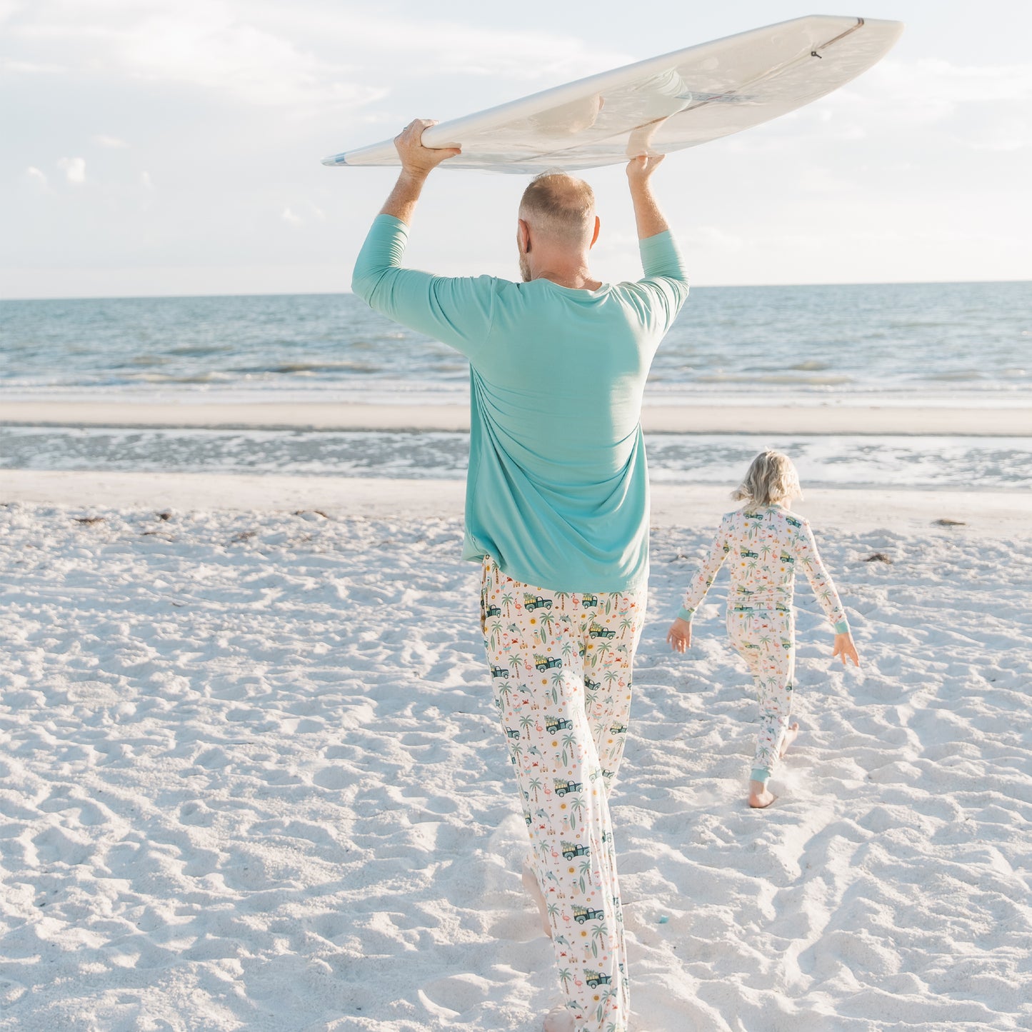 Man and child walking on a beach with a surfboard, wearing matching outfits.