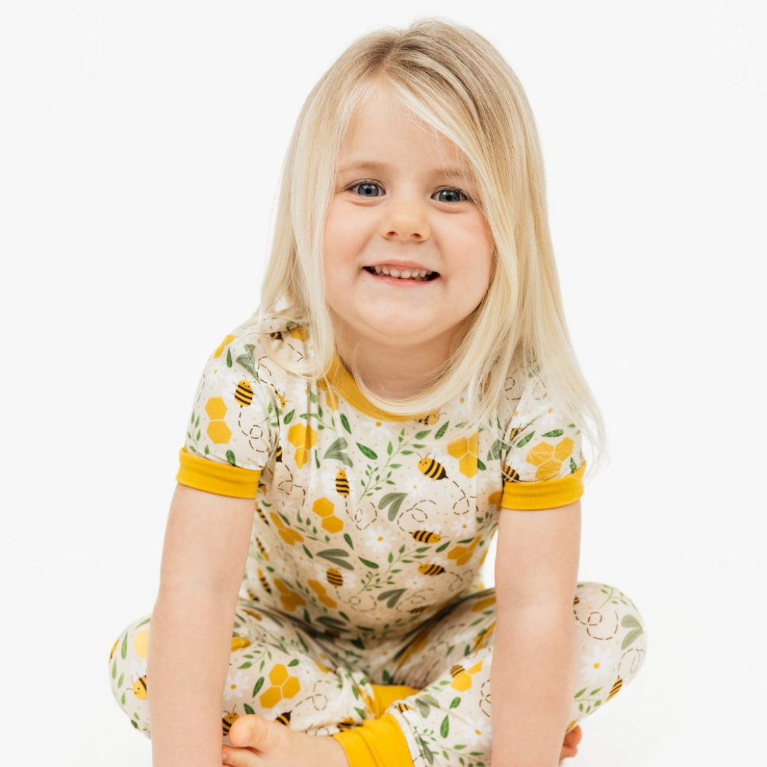 A smiling toddler girl sitting cross-legged in soft bamboo kids' pajamas with a honey bee print, designed for sensitive skin.