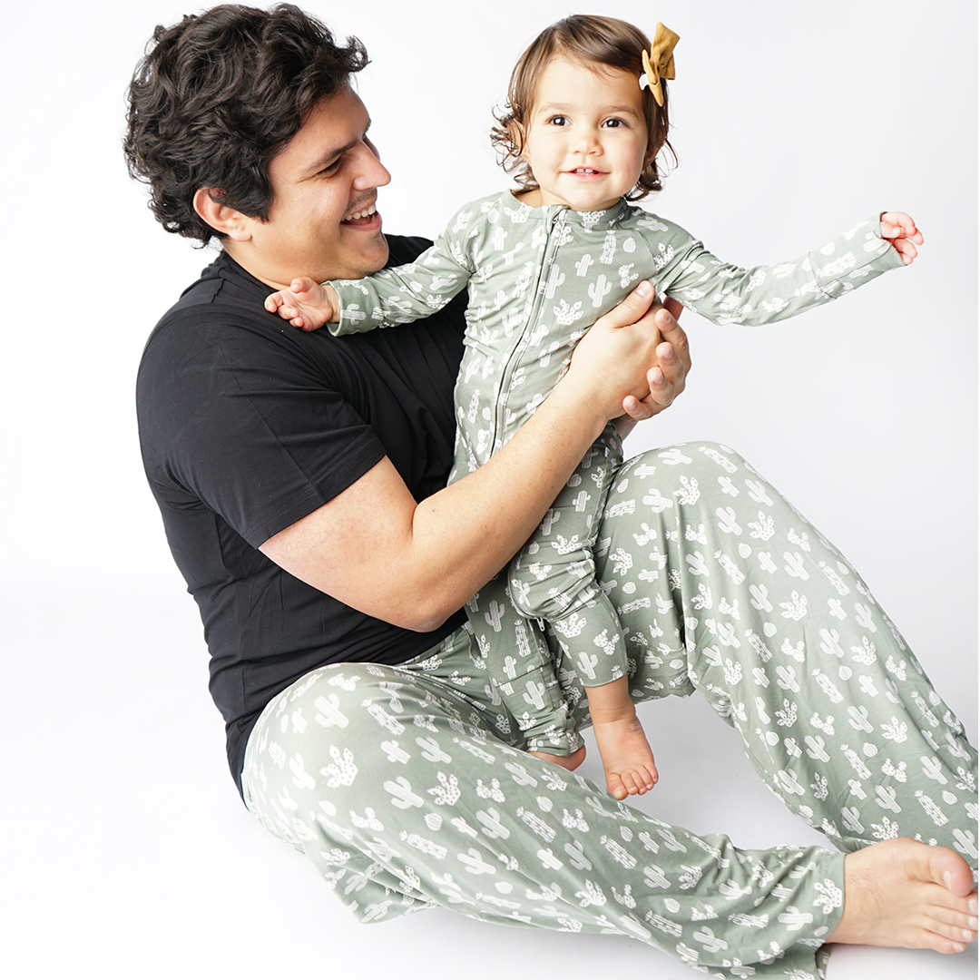 dad smiles and hold baby. he is wearing the "stay sharp" relaxed pants. the baby wears the "stay sharp" convertible. the "stay sharp" print is a variety of different white cacti on a greyish/green background. 