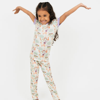 Little girl in our Magical Unicorn toddler bamboo pajama set with hands in the air. The print includes unicorns, rainbows, clouds, planets, stars, and hearts. The colors are purple, pink, and blu, with hints of green. 