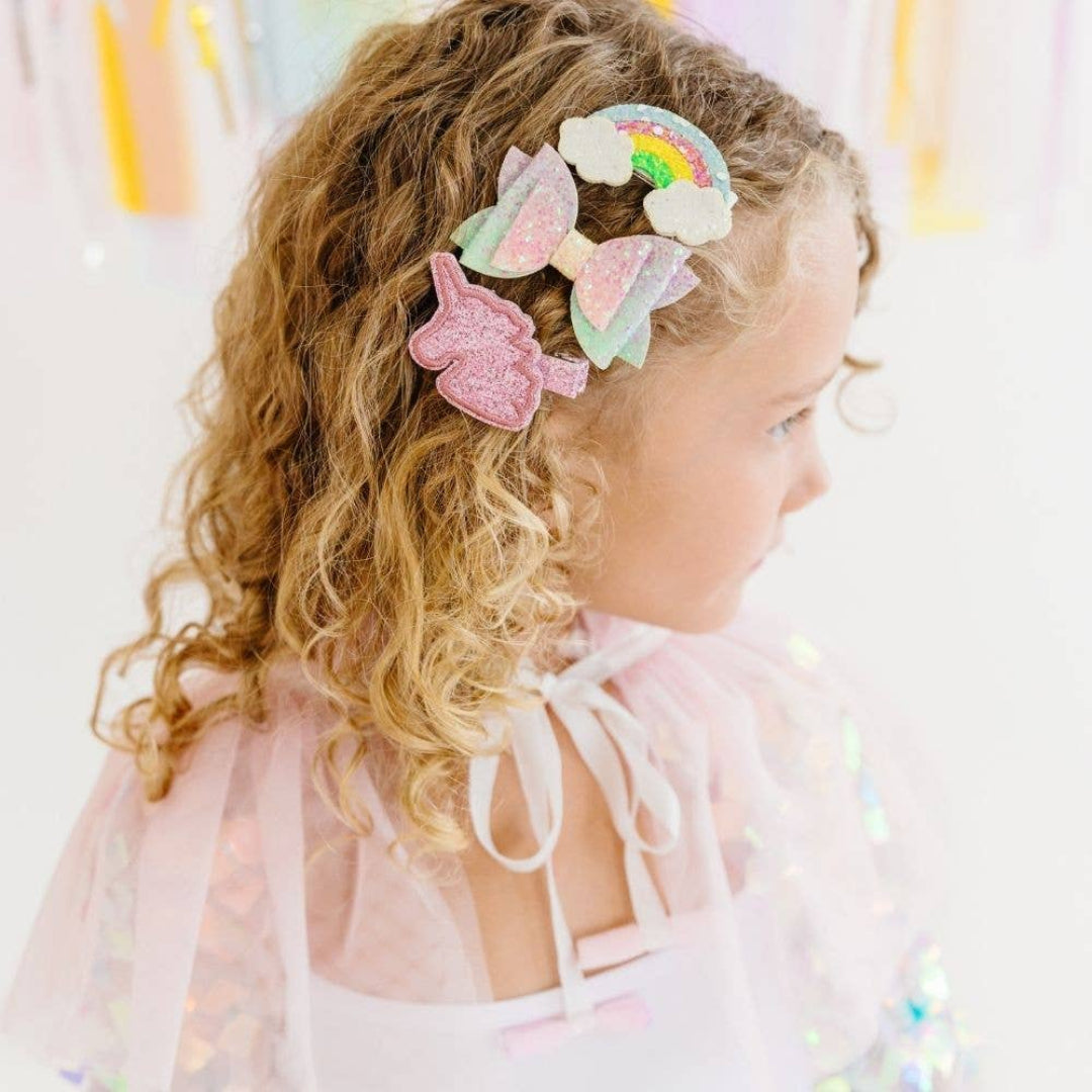 little girl wearing set of 3 glittery hair clips that include a unicorn clip, a multi colored bow, and a rainbow clip