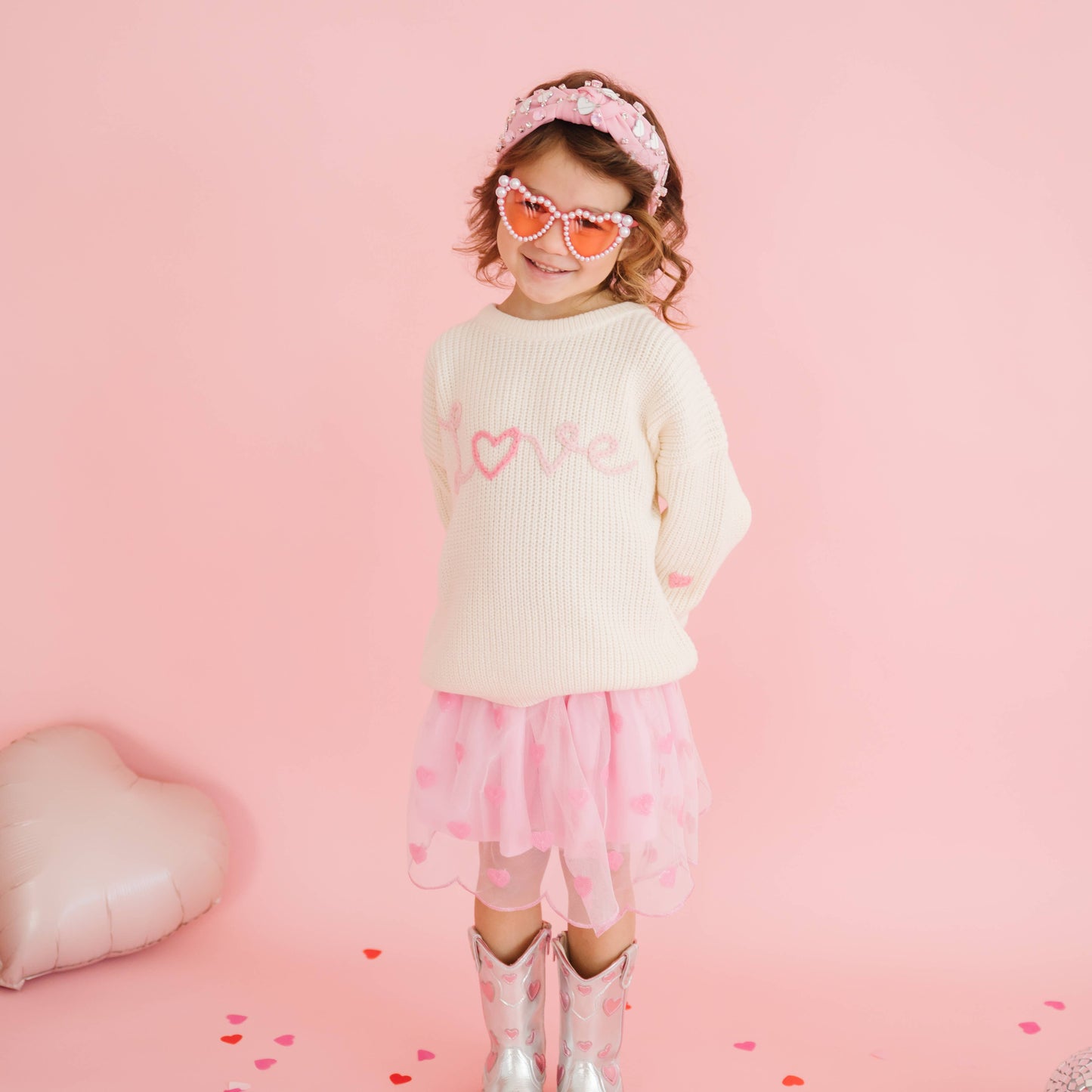 Child wearing a cream sweater with pink heart designs, pink skirt, and heart-shaped sunglasses on a pink background.