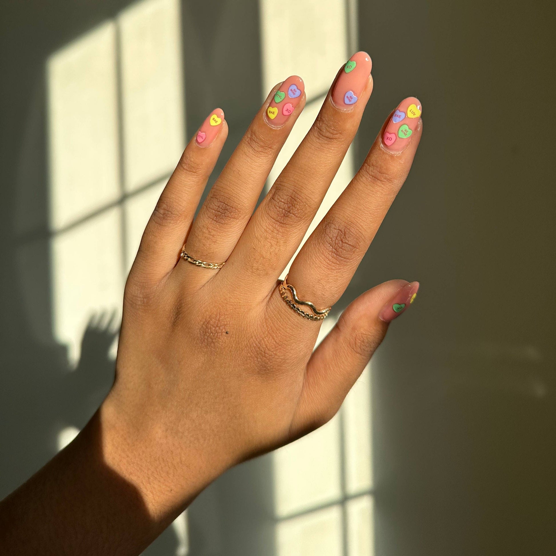 Hand with colorful nail polish and gold rings against a window with sunlight casting shadows.