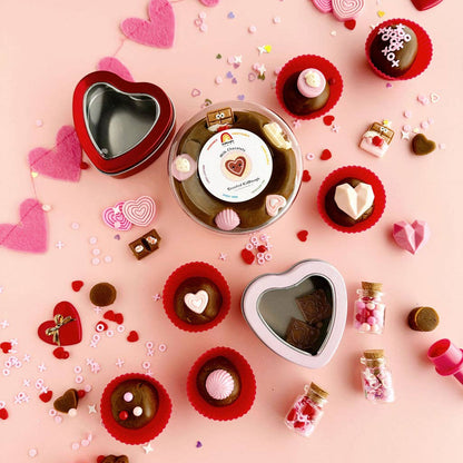 Valentine's Day-themed treats including heart-shaped boxes, candies, and small jars on a pink background.