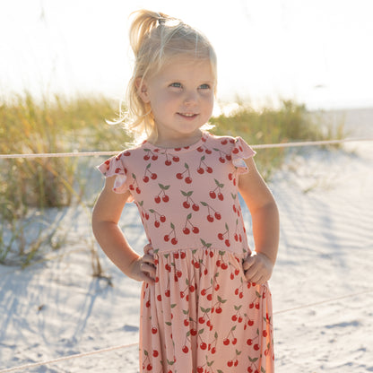 Young girl in a cherry-patterned dress standing on a sandy beach.