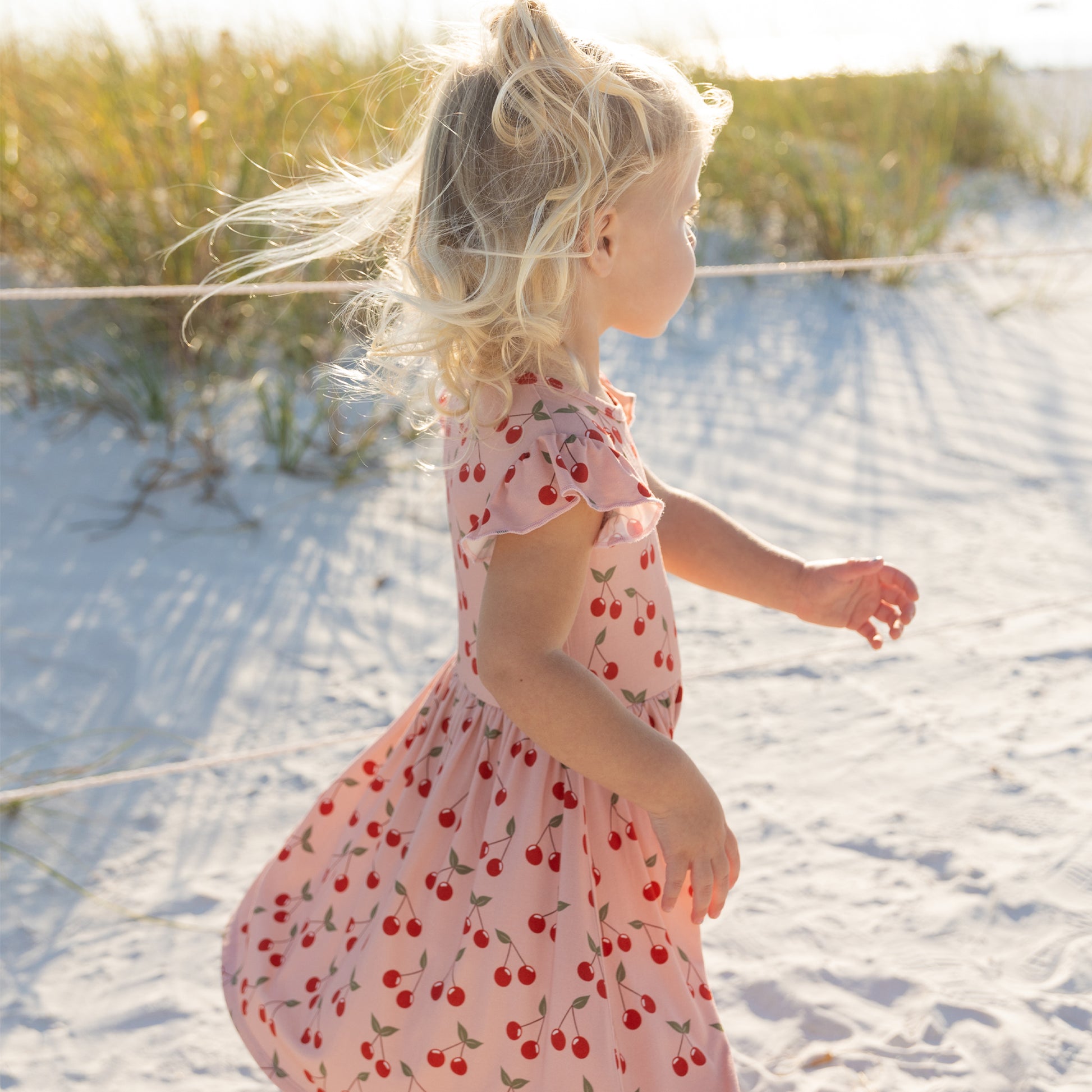 Child in a cherry-patterned dress standing on a sandy beach with grass in the background