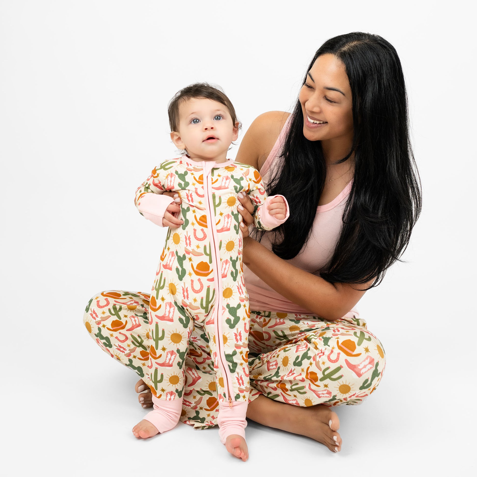 Baby girl and mom wearing our Emerson and Friends Hey Y’all Convertible Bamboo Convertible Pajamas and Hey Y'all relaxed pants. The print shows  green cactus, yellow sun, brown cowgirl hat, pink boots and horseshoes. Perfect for cozy rodeo nights or pajama parties with a cowgirl flair!