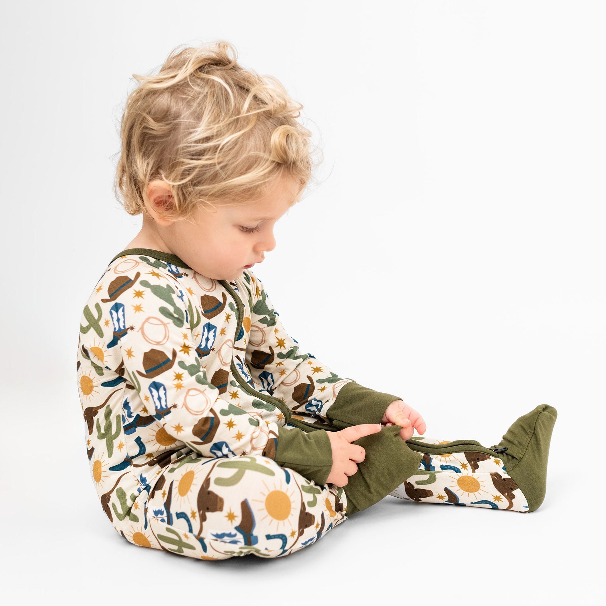 Baby boy sitting in a white background wearing  our Western Howdy Collection! This cozy bamboo convertible pajama features a charming cowboy-themed print with blue boots, cacti, suns, horseshoes, and cowboy hats. The perfect blend of comfort and western style for bedtime adventures!