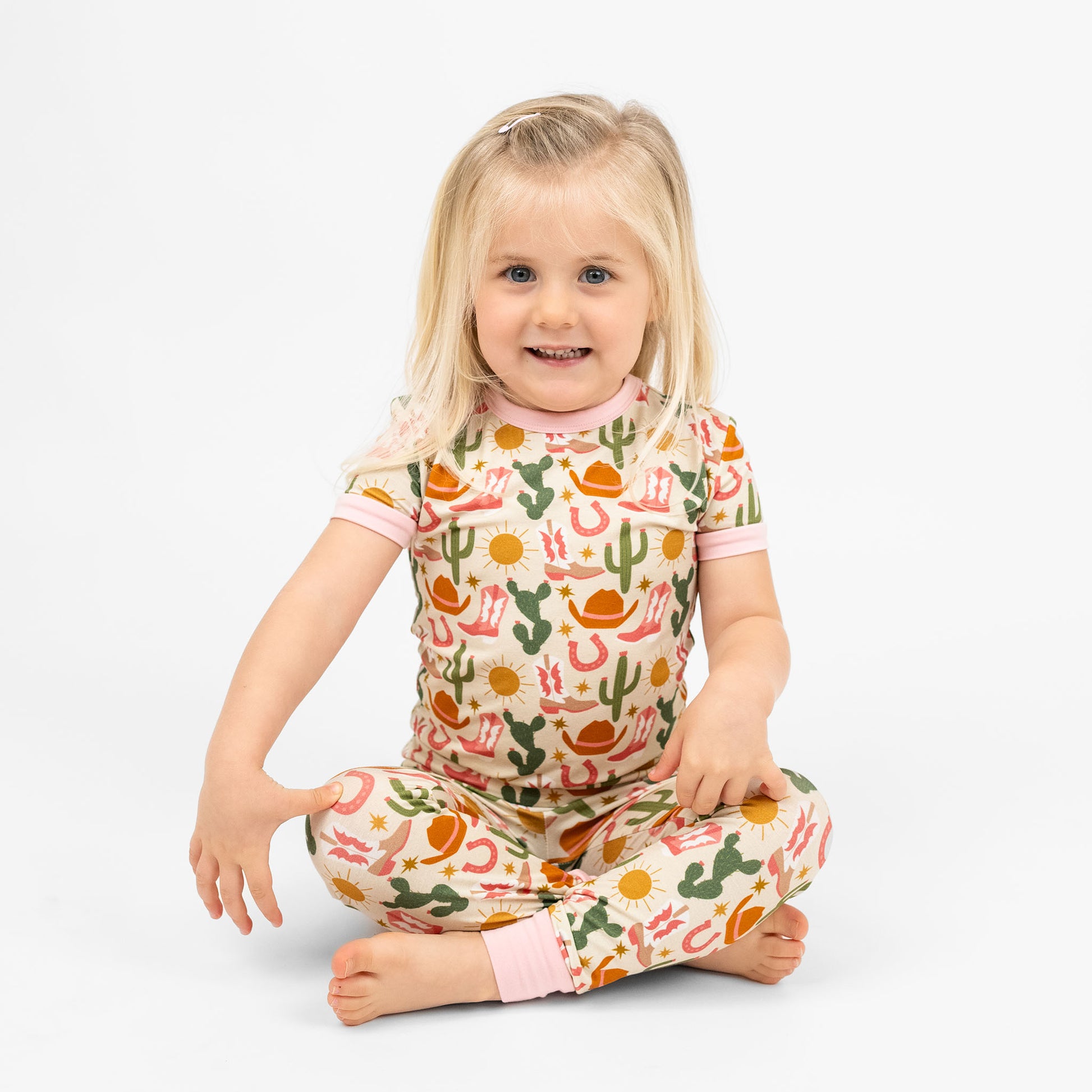 A happy girl smiling in Hey Y'all Western Bamboo Short Sleeve Kids Pajama Pants Set sitting on the floor perfect for cozy rodeo nights. 