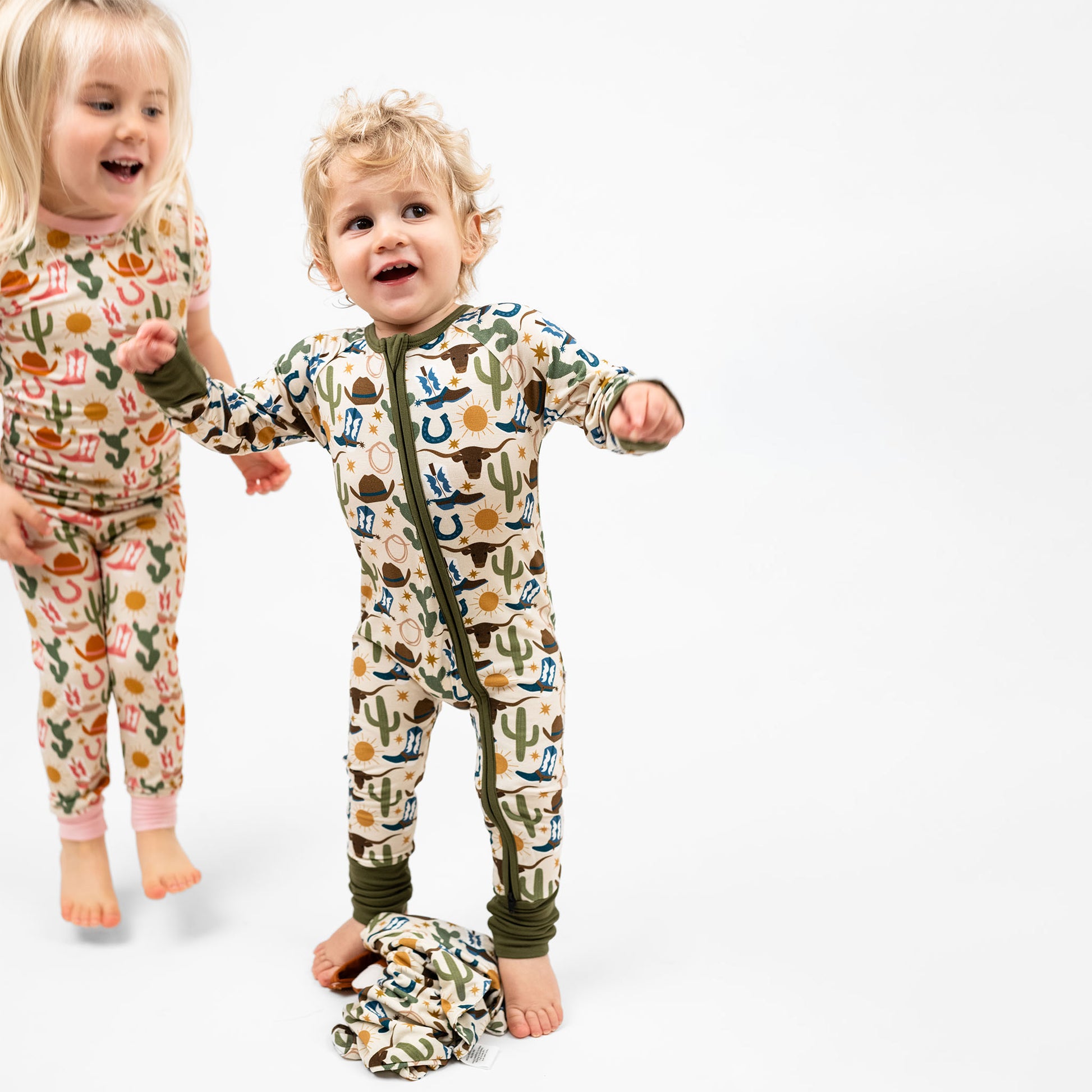 Sibling jumping happy in our Western Hey Y'all & Howdy Collection! This cozy bamboo convertible pajama features  a charming cowboy and girl-themed print with blue and pink boots, cacti, suns, horseshoes, and cowboy hats. The perfect blend of comfort and western style for bedtime adventures!