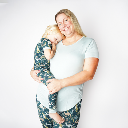 Mom and daughter pose. the mom holds her baby while the baby lays her head on her moms shoulder. the mom is wearing the "blue surf" women's top and the "dino" joggers. the baby is wearing the "dino" 2-piece matching pajama set. 